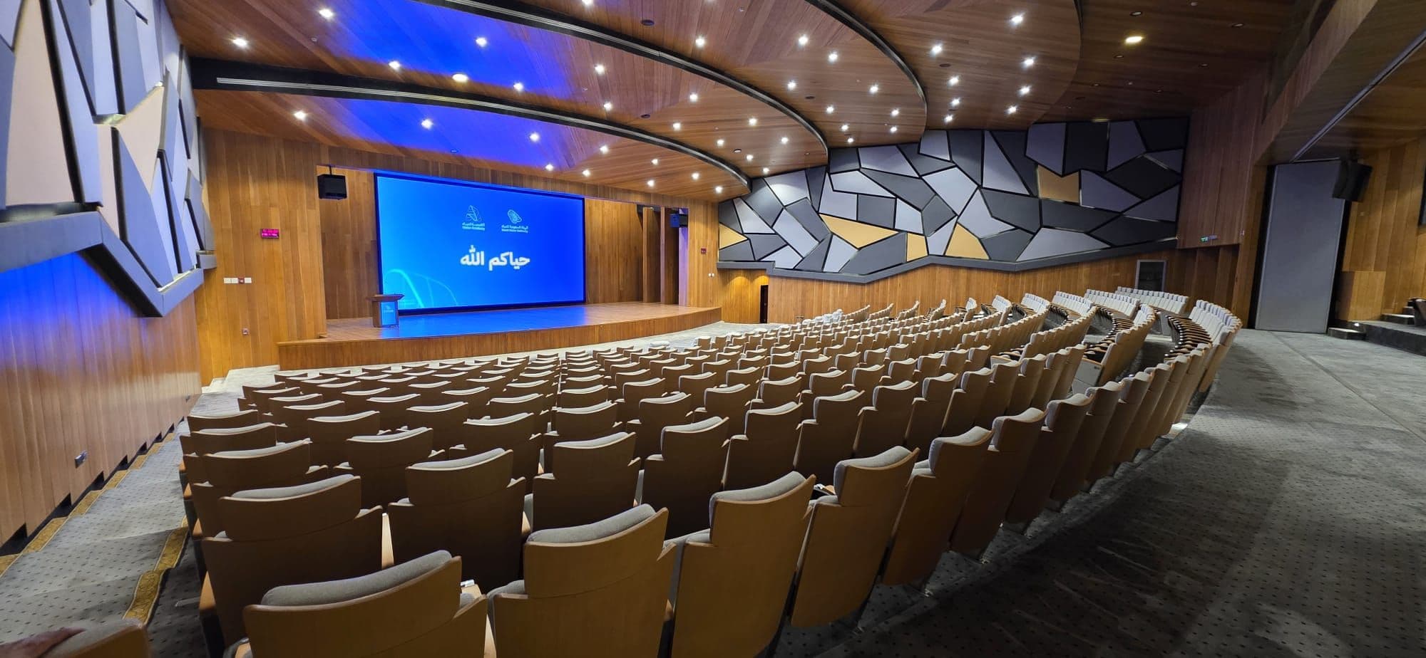 The tiered lecture theatre, timber-clad, with fixed seating facing a full-width screen.