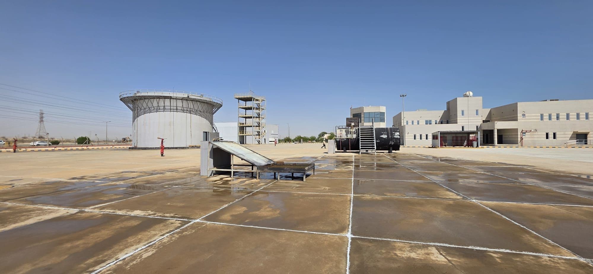 The concrete fire-ground apron seen wide, with a storage-tank prop, a drill tower and the academy's buildings behind it.