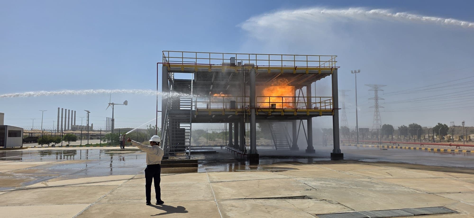 Fire burning across the first floor of the two-storey process rig on the fire ground, with a water jet playing over it and one person watching from the apron.